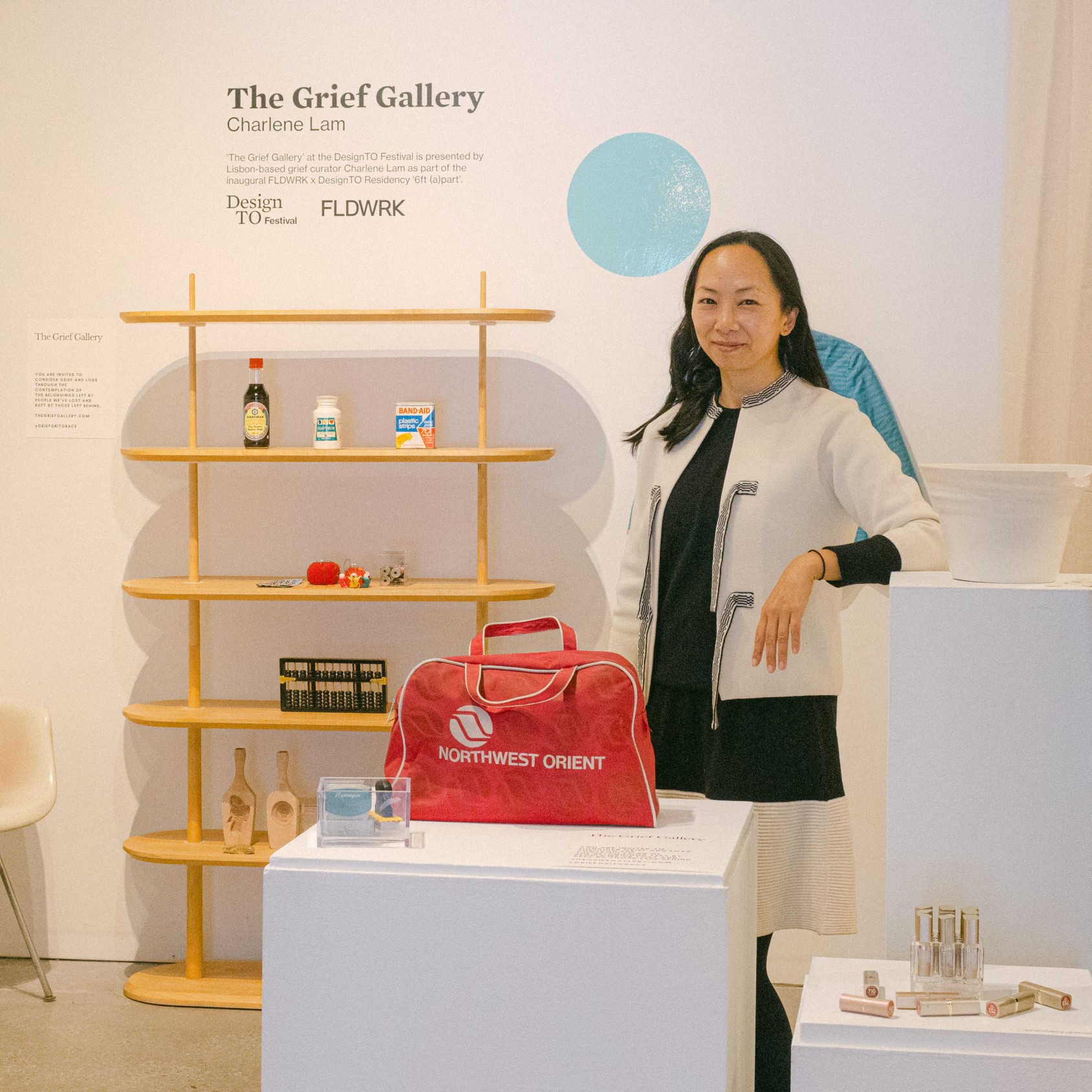 The Grief Gallery in Toronto for DesignTO, Jan 2022. Gallery space with Asian woman with white display pedestals and wooden bookcase with items displayed like vintage travel bag